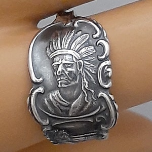 May include: A silver ring with a detailed design of a Native American man wearing a headdress. The ring is worn on a person's finger.