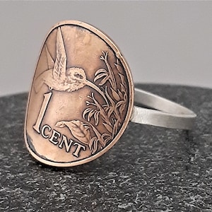 May include: A silver ring with a copper 1 cent coin featuring a hummingbird and flowers. The coin is shaped like a teardrop and the ring is plain.