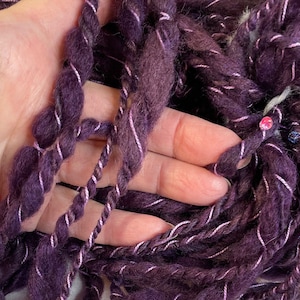 May include: A close-up of a skein of purple yarn with a white and pink stripe. The yarn is soft and fluffy, and it is perfect for knitting or crocheting.