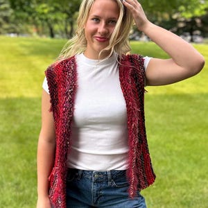 May include: A sleeveless vest in shades of red, black, and pink. The vest has a textured, fringed design. The person in the image is wearing a white t-shirt and blue jeans. The background is a green lawn and trees.