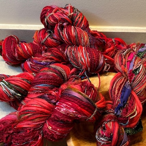 May include: A pile of red, purple, and brown yarn with a mix of textures, including some with a fuzzy look. The yarn is in a wooden bowl.