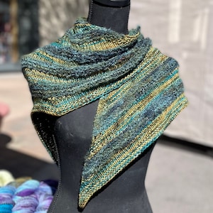 Puede incluir: Un chal triangular con un patrón de rayas en tonos verde azulado, verde y dorado. El chal se muestra en un maniquí negro.