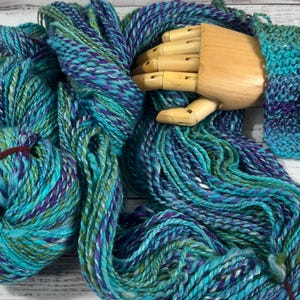 May include: A skein of hand-dyed yarn featuring a variegated color palette of turquoise, teal, deep purple, and lime green. The yarn has a thick, twisted texture and is displayed alongside a knitted swatch in the same multi-colored pattern.