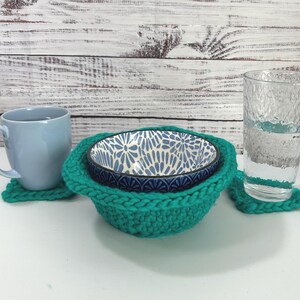 May include: A light blue mug, a clear glass, and two bowls are displayed on a white surface. The bowls are nested in a teal knitted bowl cozy. A teal knitted coaster is under the mug and glass. The bowls have a blue and white floral pattern.