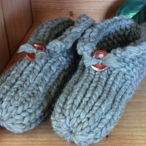 Puede incluir: Un par de pantuflas de punto gris con detalles de botones de madera. Las pantuflas están hechas de un hilo suave y peludo y tienen un aspecto cómodo y acogedor.