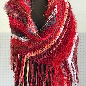 May include: A red, pink, and orange knitted shawl with a fringe border. The shawl is draped over a black mannequin.