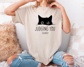 May include: A woman wearing a beige t-shirt with a black cat peeking over the top and the text "Judging You Silently". She is wearing blue jeans and sitting on a fluffy white rug.