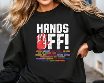 May include: Black sweatshirt with the text "HANDS OFF!" in white with a red handprint graphic. The text below the graphic lists various social and political issues in rainbow colors.