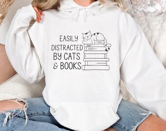 May include: White hoodie with a black and white graphic of a cat sleeping on a stack of books. The text on the hoodie reads "Easily Distracted By Cats & Books".