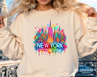 May include: A cream colored sweatshirt with a colorful graphic of the New York City skyline. The graphic features a watercolor effect with splashes of pink, blue, green, yellow, and orange. The text "NEW YORK" is in a neon yellow font.