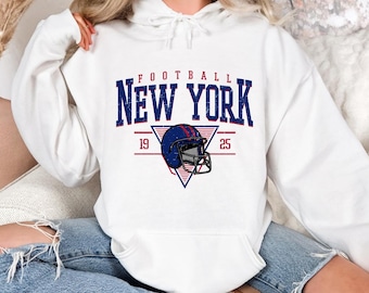 May include: White hoodie with a blue and red graphic that says "FOOTBALL NEW YORK" with a football helmet in the center and the numbers 19 and 25 on either side of the helmet.