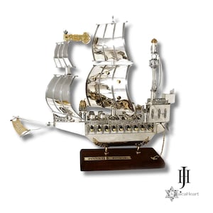 May include: A detailed silver-colored ship sculpture with intricate designs, including sails and decorative elements. The ship is mounted on a dark wooden base with Hebrew text. A gold-colored accent is visible on the ship.