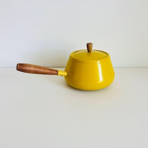 May include: A bright yellow enamelled metal saucepan with a matching lid. The pot features a long, horizontal wooden handle and a small, round wooden knob on the centre of the lid. The design is smooth, rounded, and features a vibrant yellow finish.
