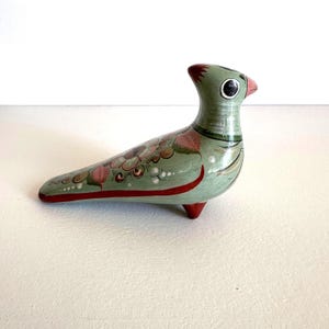 May include: A hand-painted ceramic bird figurine in shades of green, red, and pink. The bird has a long body, a small head, and is decorated with floral patterns. The figurine is resting on three small red feet.