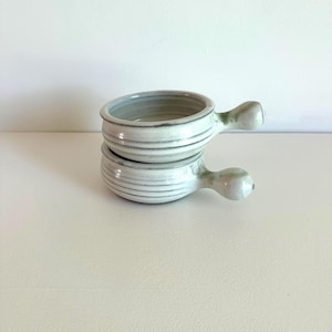 May include: Two stacked, handmade ceramic bowls with handles. The bowls are light gray with subtle green accents and horizontal ridged detailing. The handles are rounded and attached to the side of each bowl.