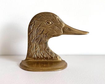 May include: A brass duck head sculpture with detailed feather etching. The sculpture is a golden color and has a long, pointed beak. The duck head is mounted on a small, rounded base, and is a decorative item.