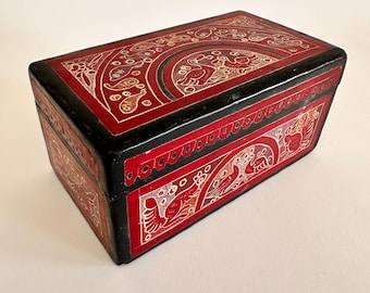 May include: A red wooden box with intricate gold and white painted designs. The box has a black border and a decorative pattern around the edge.