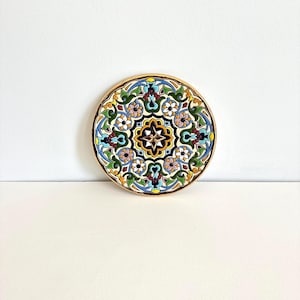 May include: A round, decorative ceramic plate with a detailed floral design. The plate features a central starburst pattern in yellow and black, surrounded by colorful flowers and leaves in shades of blue, green, and red. The rim is a warm, golden brown.