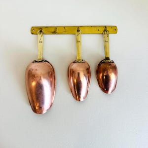 May include: A set of three copper scoops with brass handles hanging from a horizontal brass wall rack. The scoops are labelled 7oz, 3oz, and 2oz respectively. Each scoop features a rounded bowl shape and a metallic finish with visible wear.