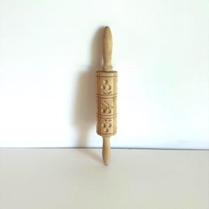 May include: A light-colored wooden rolling pin with a handle at each end. The rolling surface is decorated with a repeating cherry pattern. The rolling pin measures approximately 12 inches in length.