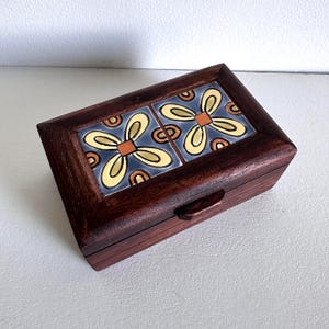 May include: A wooden box with a hinged lid and a decorative tile design on the top. The tile design features yellow, orange, and blue floral patterns.