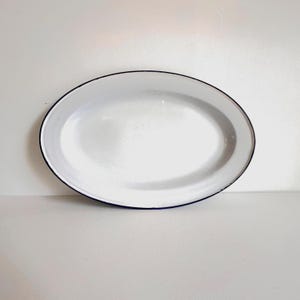 May include: Oval white enamel serving platter with a black rim. The platter is empty and sits on a white surface. The serving dish is a classic design, suitable for serving food.