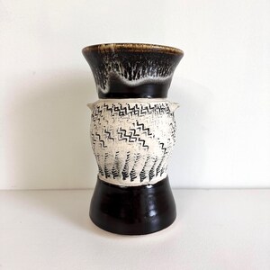 May include: A decorative ceramic vase with an hourglass shape. The top and bottom sections are dark brown, while the middle section is off-white with a black zigzag pattern. The rim has a textured, dripped glaze.