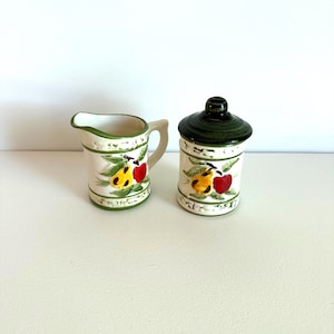 May include: A cream-colored ceramic creamer and sugar bowl set. The set features a green rim and is decorated with yellow pears, red apples, and green leaves. The sugar bowl has a dark green lid.