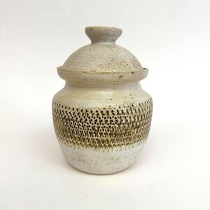 May include: A ceramic jar with a lid. The jar is off-white with a band of brown speckled dots around the middle. The lid is a similar colour and texture, with a small knob on top.