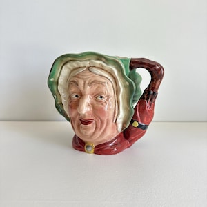 May include: A ceramic mug shaped like an elderly woman's head. The mug features a detailed face with wrinkles, a white wig, and a green and white bonnet. The handle is a dark brown color, and the woman is wearing a red garment with a yellow and black accent.