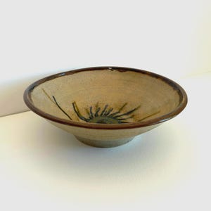 May include: A handmade ceramic bowl with a beige exterior and a dark brown rim. The interior features a sunburst design in shades of green and blue. The bowl is a unique piece of pottery.