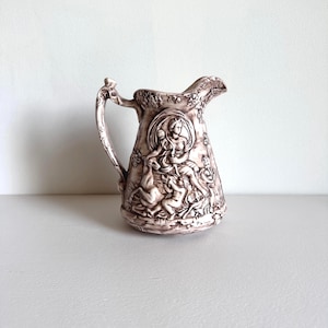 May include: A white ceramic pitcher with a brown relief design of a woman and figures. The pitcher has a handle and a spout.