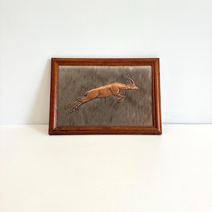 May include: A framed artwork depicting a leaping antelope. The antelope is a warm brown hue, set against a textured grey background. The frame is dark wood. The artwork is displayed on a white surface.