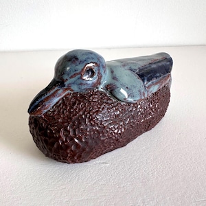May include: A ceramic duck figurine with a textured, dark brown body and a blue-grey head and back. The duck has a detailed beak and eye, and the glaze gives it a glossy finish. The figurine is a decorative item.