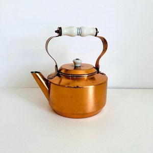 May include: A copper tea kettle with a cylindrical body and a short spout. The kettle features a curved metal handle topped with a white ceramic grip accented by blue stripes. A matching white ceramic knob sits on the centre of the lid.