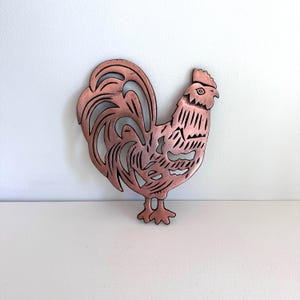 May include: A copper-coloured metal rooster cutout. The rooster features an intricate design with cut-out sections in its body, tail, and comb. The rooster is positioned on a white surface, with a white wall in the background.