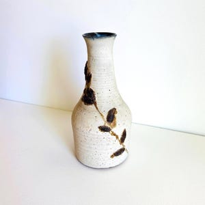 May include: A tall, off-white ceramic vase with a speckled texture and a narrow neck. The exterior features a dark brown, embossed branch design with small, textured leaves extending vertically along the side. The rim has a dark blue accent.