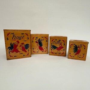 May include: Set of four wooden canisters with rooster illustrations. The canisters are labeled "Flour", "Sugar", "Coffee", and "Tea".
