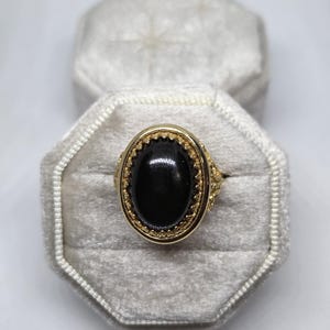May include: A gold-toned ring with an oval, black gemstone. The ring is set in a detailed gold frame and rests inside a gray velvet ring box. The ring's design features intricate detailing around the gemstone.