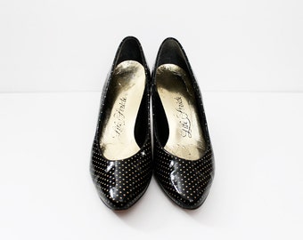 May include: Pair of black patent leather pumps with gold polka dots. The shoes have a pointed toe and a low heel. The insole is gold and has the text "Lila Freida" printed on it.