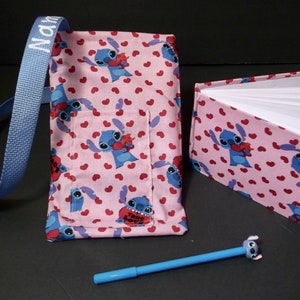 May include: A pink fabric tote bag with a blue Stitch character holding a red heart. The bag has a blue strap and a pocket on the front. A matching pink fabric notebook with Stitch on the cover is open to reveal blank pages. A blue pen with a Stitch character on the end is also included.