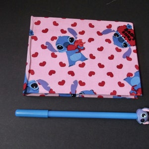 May include: A pink fabric notebook with a repeating pattern of blue Stitch characters holding hearts and red hearts. The notebook has the text "Back 2 School" on the cover.