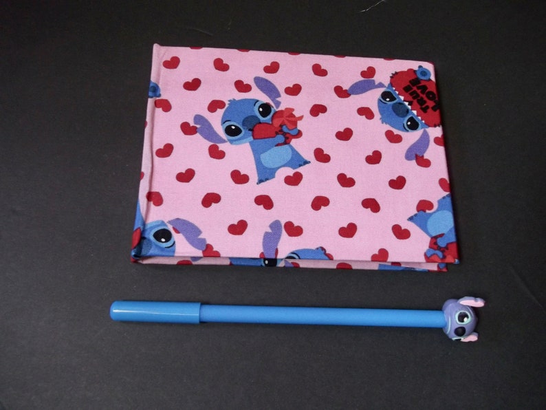 May include: A pink fabric notebook with a repeating pattern of blue Stitch characters holding hearts and red hearts. The notebook has the text "Back 2 School" on the cover.