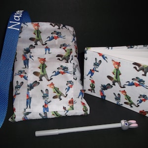 Peut inclure: Un sac en tissu blanc, un carnet et un stylo avec des personnages de dessins anim&eacute;s. Le sac a une sangle bleue avec le nom "Name" brod&eacute; dessus. Le carnet est ouvert, r&eacute;v&eacute;lant des pages vierges. Le stylo a un dessus en forme de lapin.