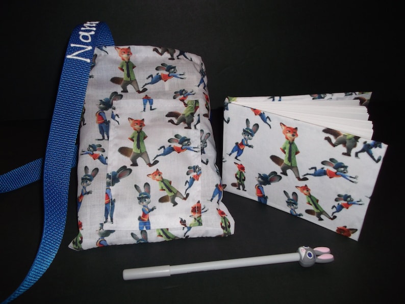 Peut inclure: Un sac en tissu blanc, un carnet et un stylo avec des personnages de dessins anim&eacute;s. Le sac a une sangle bleue avec le nom "Name" brod&eacute; dessus. Le carnet est ouvert, r&eacute;v&eacute;lant des pages vierges. Le stylo a un dessus en forme de lapin.