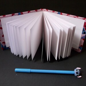 May include: A pink and blue fabric-covered notebook with white pages is open to reveal the blank pages. A blue pen with a blue cartoon character on the end is lying on a black surface.