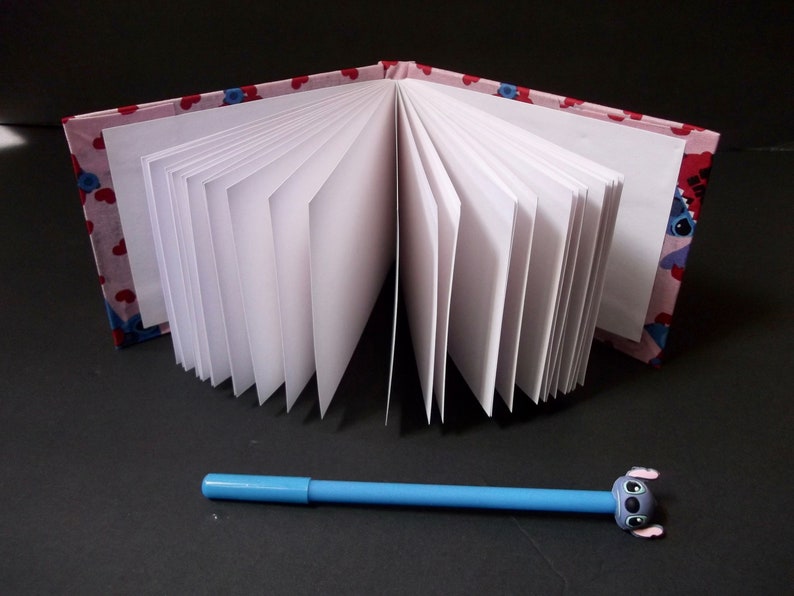 May include: A pink and blue fabric-covered notebook with white pages is open to reveal the blank pages. A blue pen with a blue cartoon character on the end is lying on a black surface.