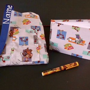May include: A white fabric bag with a blue strap and a pocket. The bag has a repeating pattern of Toy Story characters and the words "Toys at Play". A small white notebook with the same pattern is next to the bag. A yellow pen is on the table.