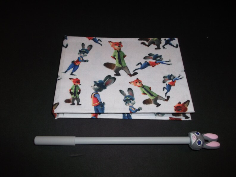 Peut inclure: Un tissu blanc pli&eacute; avec un motif r&eacute;p&eacute;t&eacute; de personnages de dessins anim&eacute;s de renards et de lapins. Un stylo gris avec un dessus en forme de lapin est devant. Les personnages sont dans diverses poses et tenues.