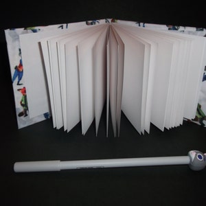 Peut inclure: Un cahier ouvert avec des pages blanches et une couverture &agrave; motifs avec des personnages de dessins anim&eacute;s color&eacute;s. Un stylo blanc avec un dessus en forme de lapin gris repose devant le cahier sur une surface noire.